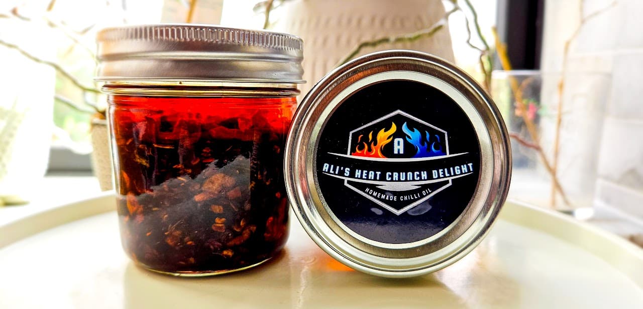 A jar of Ali's Heat Crunch Delight chilli oil next to its lid, which shows the AHCD logo label.