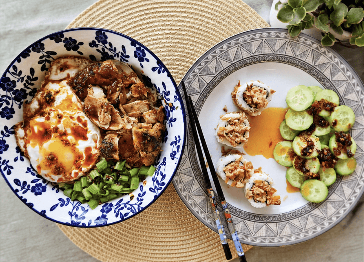 An overhead shot of two dishes: a noodle bowl topped with a fried egg, shredded chicken and chilli oil, beside a plate of sushi rolls and cucumber slices drizzled with chilli oil.